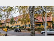 Reihenhäuser in einem Wohnviertel mit gelb gestrichenen Fassaden, roten Ziegeldächern und grünen Fensterläden. Im Vordergrund stehen Bäume, Straßenschilder, ein Briefkasten und parkende Autos entlang einer ruhigen Straße.