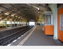 Bahnhofshalle eines S-Bahnhofs mit überdachten Bahnsteigen, gewölbter Stahlkonstruktion und Gleisen in der Mitte. Die Wände sind teilweise mit orangefarbenen Fliesen verkleidet, und es gibt Hinweisschilder sowie ein kleines Fahrkartenhäuschen an der Seite.
