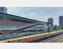 Außenansicht eines modernen Einkaufszentrums mit der Aufschrift "East Side Mall", im Vordergrund verlaufen Bahngleise mit einem vorbeifahrenden Zug der Berliner S-Bahn. Im Hintergrund sind hohe Gebäude und ein Kran zu sehen.