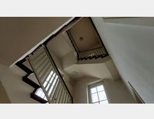 Treppenhaus mit mehreren Wendungen, weiß verputzten Wänden und Holzgeländern; eine rechteckige Fensteröffnung mit Gitterverstrebung lässt Tageslicht herein.