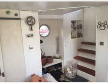 Innenansicht eines kleinen, weißen Raumes mit einer Holztreppe. Ein rundes Bullauge, Regalfächer an der Wand und verschiedene Gegenstände wie Blumentöpfe und Schuhe prägen den Raum.