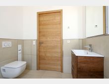 Modernes Badezimmer mit hellen Wand- und Bodenfliesen, einer Holztür, einem wandhängenden WC sowie einem Waschbecken mit Holzunterschrank und Wandspiegel.