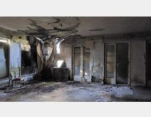 Ein verlassener und heruntergekommener Raum mit beschädigten Wänden und Decke, freiliegender Isolierung und zerbrochenen Fensterläden. Der Boden ist mit Schutt und Staub bedeckt, eine einzelne alte Heizung und ein Stuhl befinden sich am Rand des Raumes.