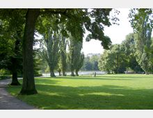 Grüne Parklandschaft mit großen Bäumen und einer offenen Rasenfläche, im Hintergrund ein See und vereinzelte Spaziergänger. Ein gepflasterter Weg verläuft links durch den schattigen Bereich des Parks.