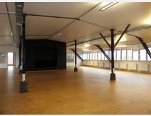 Geräumiger, lichtdurchfluteter Raum mit Holzfußboden, schwarzen Stahlträgern zur Deckenkonstruktion und einer großen Fensterfront auf der rechten Seite; im Hintergrund eine schwarze Wand mit rechteckiger Nische.