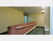 Ein Korridor mit schräger Rampe, flankiert von einer niedrigen Ziegelmauer und einer Wand, beide mit weißen Handläufen ausgestattet. Am Ende der Rampe befindet sich eine Glastür mit der Aufschrift "Ausgang".