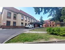 Eingangsbereich einer Notaufnahme mit einem hellen zweistöckigen Gebäude aus sandfarbenem Stein und roten Ziegeln. Im Vordergrund ist eine Zufahrt mit einem geparkten weißen Fahrzeug, daneben Grünflächen und eine Bushaltestelle.