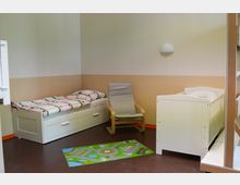 Kinderzimmer mit heller Einrichtung: Links ein Einzelbett mit Bettkasten und bunter Bettwäsche, daneben ein grauer Schaukelstuhl. Rechts ein weißes Gitterbett vor einer zweifarbigen Wand in Beige und Creme. Auf dem braunen Boden liegt ein kleiner, bunter Spielteppich mit Straßenmuster.