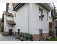 Seitenansicht eines Wohnhauses mit weiß verputzter Fassade, rotem Ziegeldach und Ziegelsteinsockel. Es gibt vergitterte Fenster, ein kleines Vordach mit Dachrinnen und eine Gartenfläche mit Pflanzen im Vordergrund.