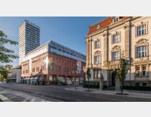 Straßenansicht mit moderner Architektur und historisch inspiriertem Gebäude: Links ein mehrstöckiger, moderner Glas- und Backsteinbau, im Hintergrund ein hoher Büroturm mit der Aufschrift „Der Oderturm“. Rechts ein repräsentatives, denkmalgeschütztes Gebäude mit verzierten Fassadenelementen und einem roten Dach.