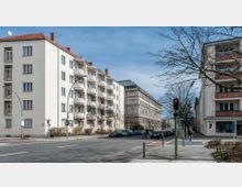 Straßenszene in einer Stadt mit mehrstöckigen Wohngebäuden im Vordergrund, eines davon mit runden Balkonen. Im Hintergrund steht ein großes, steinernes Gebäude im klassischen Architekturstil. Am Straßenrand sind Autos geparkt, und eine Fußgängerampel zeigt rot.