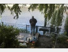 Ein Mann steht an einem Zaun am Ufer eines Flusses und angelt. Um ihn herum befinden sich Büsche, ein schattenspendender Baum und einige persönliche Gegenstände wie eine Thermoskanne und Taschen auf einer Mauer.