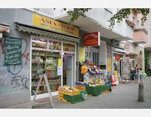 Straßenansicht eines kleinen Asia-Lebensmittelgeschäfts mit gelbem Schild, das Obst, Gemüse und Getränke anbietet. Vor dem Laden stehen Obst- und Gemüsekisten sowie Aufsteller mit Angeboten, daneben befindet sich ein Zeitschriftenladen mit Zeitungsständern am Gehweg.