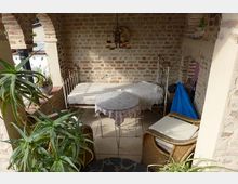 Gemütlicher, überdachter Sitzbereich mit verputzten Ziegelwänden und offenen Rundbögen. Der Raum enthält ein Metallbett, einen kleinen runden Tisch mit Tischdecke, geflochtene Sessel und Pflanzen.