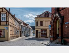 Straßenszene in einer Altstadt mit Fachwerkhäusern auf der linken Seite, einem hellgelben Gebäude mit abgerundeter Ecke in der Mitte und einem gemauerten roten Gebäude rechts. Kopfsteinpflaster bedeckt die Straße, und verschiedene Straßenschilder sind sichtbar.