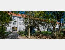 Mehrfamilienhaus mit weißer Fassade und rotem Ziegeldach, das durch einen Torbogen in einen Innenhof führt; im Vordergrund ein gepflasterter Weg, Bäume und ein Maschendrahtzaun.