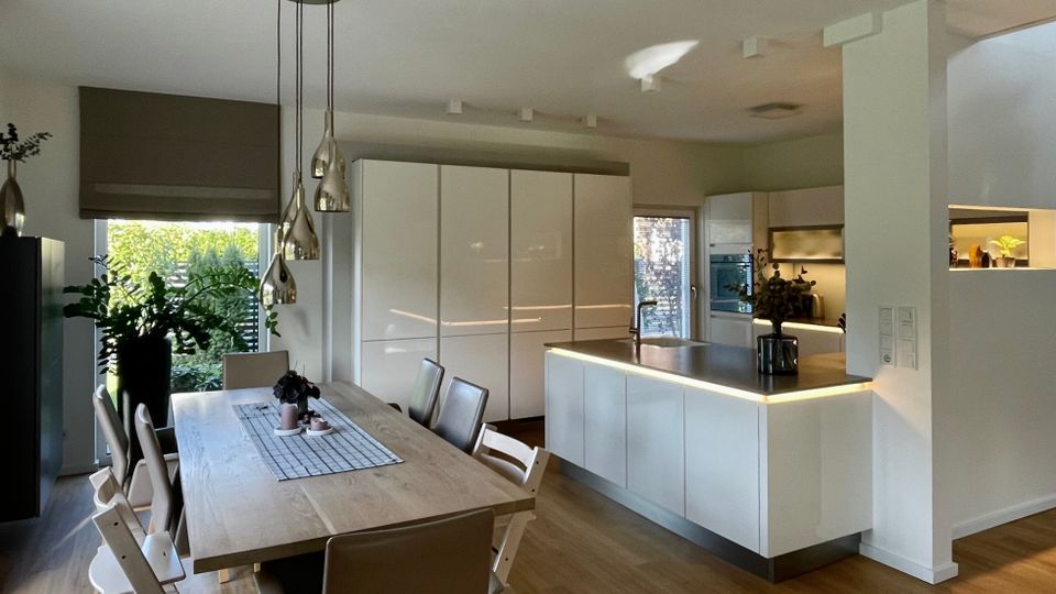 Offener Wohn- und Essbereich mit moderner, weißer Einbauküche und Kücheninsel mit dunkler Arbeitsplatte sowie indirekter Beleuchtung. Daneben steht ein großer Holzesstisch mit Stühlen unter hängenden Pendelleuchten, mit großen Fenstern und Blick ins Grüne.