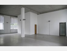 Großer leerer Raum mit weißen Wänden, grauem Betonboden und einer freiliegenden Betondecke. Der Raum hat große Fenster mit Jalousien, eine Holzinnentür und eine Tür mit mattierter Glasscheibe.