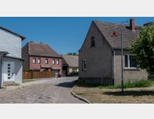 Straßenszene in einem Dorf mit Kopfsteinpflaster, umgeben von einfachen Wohnhäusern mit Satteldächern. Im Hintergrund steht ein rotes Backsteingebäude mit der Aufschrift „Fleischerei Reihs“, daneben Bäume und ein klarer blauer Himmel.