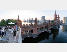 Blick auf die Oberbaumbrücke in Berlin bei Sonnenuntergang, mit ihren markanten roten Backsteintürmen und Bögen. Im Vordergrund befindet sich eine Terrasse mit Menschen, die an Stehtischen und Sitzgelegenheiten stehen und den Ausblick genießen, dahinter die Spree und moderne Gebäude.