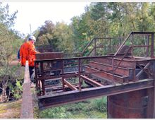 Eine Person in Arbeitskleidung und Schutzhelm steht auf einer alten, rostigen Stahlkonstruktion in einer bewaldeten Umgebung. Im Hintergrund sind dichte Bäume und ein klarer Himmel sichtbar.
