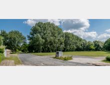 Eine ruhige Parklandschaft mit einer großen Wiese und hohen, dichten Bäumen im Hintergrund. Im Vordergrund verlaufen ein asphaltierter Weg, ein kleiner Verteilerkasten sowie ein Straßenschild, und links sieht man einige Garagen.