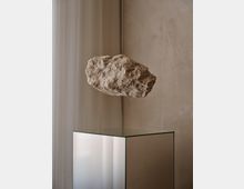 Innenraum mit minimalistischer Präsentation: Ein schwebender, rauer Steinblock hängt über einem würfelförmigen Sockel aus spiegelndem Glas. Im Hintergrund sind eine beige Wand und ein halbtransparenter Vorhang zu sehen.