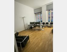 Büroeinrichtung mit hellem Holzboden, einem Schreibtisch mit mehreren Monitoren, einer Stehlampe und zwei Fenstern mit Jalousien. Im Raum stehen außerdem schwarze Stühle und ein Sessel mit Metallrahmen.