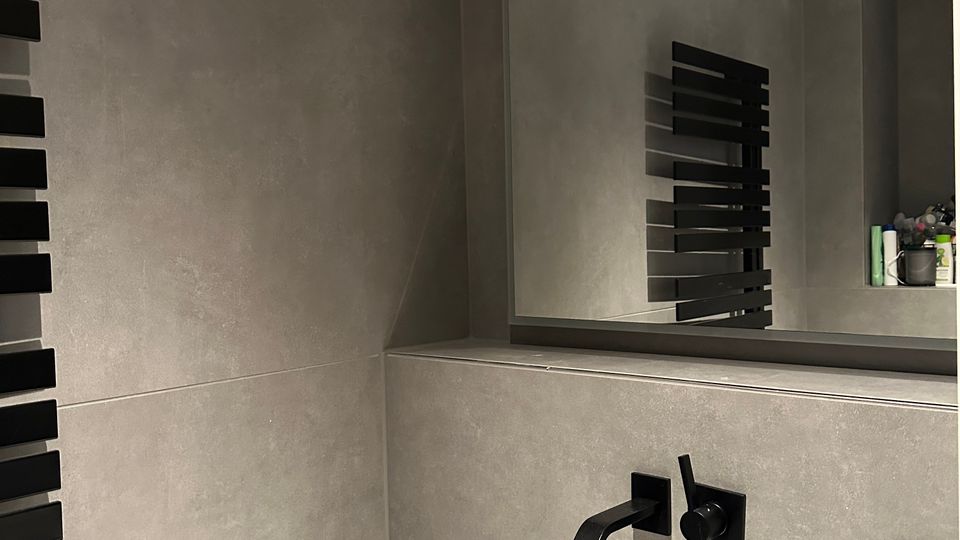 Modernes Badezimmer mit grauen Wandfliesen, rechteckigem weißem Waschbecken und mattschwarzer Wandarmatur. Darüber hängt ein großer Spiegel mit dunklem Rahmen; am Rand ist ein schwarzer Handtuchheizkörper zu sehen.