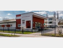 Außenansicht eines Amtsgerichtsgebäudes mit roter und weißer Fassade, der Hausnummer 58 und einer großen Eingangstür. Im Vordergrund ein schwarzer Zaun sowie blühende Büsche, im Hintergrund weitere moderne Gebäude und Bäume.