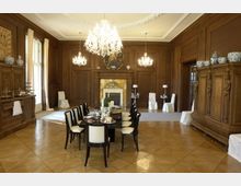 Großer, eleganter Speisesaal mit Holzvertäfelung und aufwendigen Kronleuchtern. Ein runder Esstisch mit schwarzen Stühlen steht im Zentrum, während eine Kaminverkleidung, Schränke und dekorative Vasen den Raum schmücken.