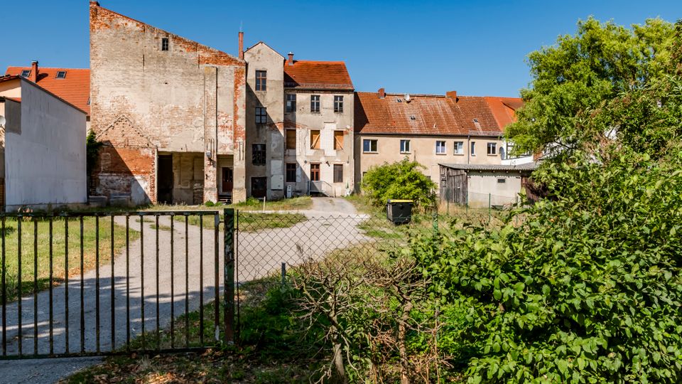 Blick durch ein Metalltor in einen kleinen Hinterhof mit Kiesweg und Grasflächen, umgeben von älteren, teils verputzten Gebäuden mit roten Ziegeldächern und verwitterten Fassaden. Rechts begrenzen dichter Bewuchs und Bäume das Gelände, daneben stehen ein kleiner Schuppen und Nebengebäude.