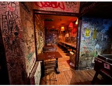Schmaler Innenraum einer Bar oder eines Cafés mit Holzboden, einfachen Tischen und Hockern entlang einer langen Theke. Wände, Türrahmen und Decke sind dicht mit Graffiti und Aufklebern bedeckt, beleuchtet von warmen, hängenden Lampenschirmen.