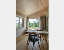 Heller Raum mit Holzdecke und holzfarbenem Fußboden, eingerichtet mit einem schlichten Holztisch und schwarzen Stühlen. Große Fensterfront mit Blick auf eine grüne Landschaft, Vorhänge und Sitzkissen auf einer Fensterbank im Hintergrund.