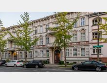 Straßenansicht eines historischen, mehrstöckigen Wohnhauses mit reich verzierten Fassadenelementen und großen Fenstern. Vor dem Gebäude stehen Bäume und parkende Autos entlang einer Straße mit Fußweg.
