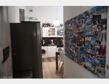 Enger Wohnungsflur mit Blick in eine kleine Küche, ausgestattet mit weißen Schränken, einer Waschmaschine und einer Arbeitsfläche mit Kaffeezubereiter. Rechts an der Wand ist eine große Fotocollage mit vielen Bildern angebracht.