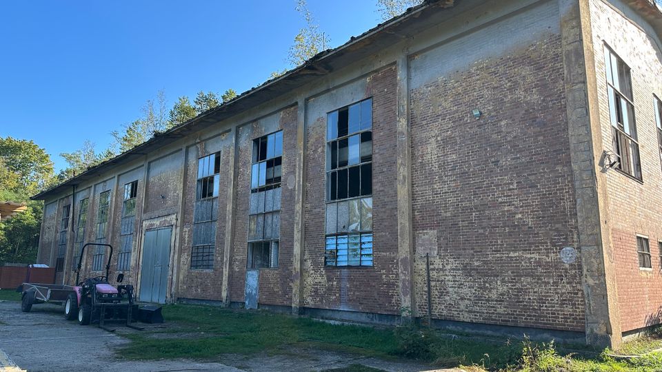 Langes, verlassen wirkendes Industriegebäude aus Backstein mit Betonpfeilern und großen, teils zerbrochenen Fensterflächen entlang der Fassade. Davor liegen ein betonierter Hof und etwas Gras, im Hintergrund sind Bäume unter blauem Himmel zu sehen.