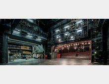 Ein Blick hinter die Bühne eines Theaters mit schwarzer Bühnenfläche, offenen technischen Konstruktionen und zahlreichen Scheinwerfern. Links sind Lagerflächen mit gestapeltem Bühnenequipment und rechts der leere Zuschauerraum mit ansteigenden Sitzreihen sichtbar.