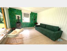 Wohnraum mit heller Holzoptik auf dem Boden und einer grünen Akzentwand mit tropischem Muster. Der Raum verfügt über einen Kaminofen, ein grünes Sofa, einen weißen Schaukelstuhl mit Fellbezug und ein großes Fenster, das Tageslicht hereinlässt und den Blick ins Freie freigibt.