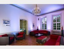 Großzügiger, stilvoller Raum mit Holzparkettboden, weißen Wänden und großen Fenstern. Der Raum ist mit modernen schwarzen Rattanmöbeln mit roten Polstern eingerichtet, besitzt einen Kronleuchter an der Decke und einen Durchgang zu einem angrenzenden, prunkvoll dekorierten Esszimmer.