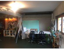 Arbeitsbereich in einem Atelier mit industriellem Stil: Zu sehen sind ein großer Tisch mit zwei Bürostühlen, Staffeleien, Regale mit Materialien sowie eine Zimmerpflanze. An der Wand hängt ein abstraktes Kunstwerk, und ein Fenster auf der rechten Seite lässt Tageslicht herein.