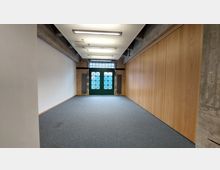 Leerer Raum mit Teppichboden, links eine weiße Wand, rechts eine Wand aus Holzpaneelen und im Hintergrund eine grüne Doppeltür mit Gittermuster und darüber Glasbausteine. Die Decke zeigt freiliegende Balken und moderne Hängelampen.