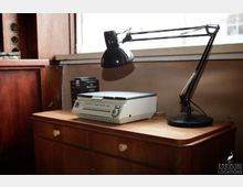 Ein Holzschreibtisch mit einer Tischlampe, einem alten Kompaktkopierer und Büchern; im Hintergrund ein Fenster und eine hölzerne Wandverkleidung.