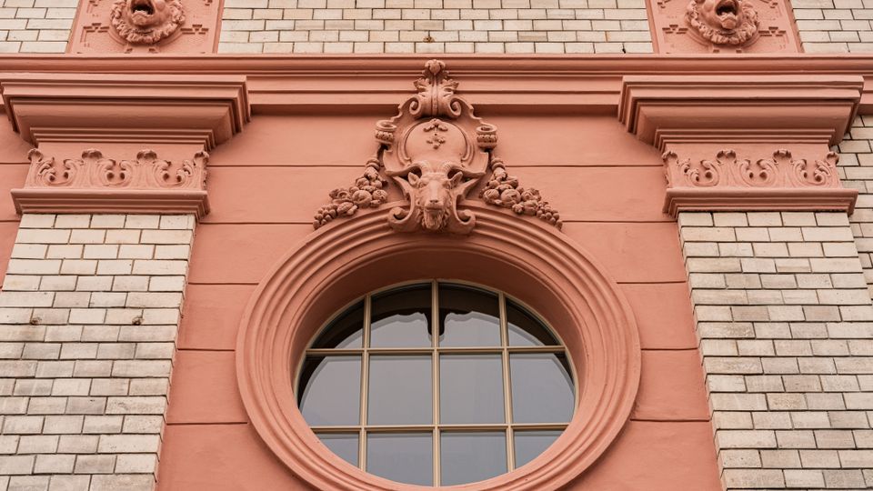 Nahaufnahme einer historischen Backsteinfassade mit rosafarbenen Stuckelementen und einem runden Fenster mit Sprossen. Über dem Fenster befindet sich ein reich verziertes Ornament mit Maskenrelief und floralen Details.