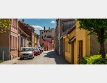 Schmale gepflasterte Straße in einem Wohngebiet mit pastellfarbenen Häusern auf beiden Seiten; geparkte Autos reihen sich entlang der linken Straßenseite, während im Vordergrund ein Baum und Sträucher zu sehen sind. Der Himmel ist teils bewölkt und blau.