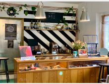 Ein kleines Café mit einer Holztheke, die Schmuck und andere Gegenstände im Glasdisplay ausstellt. Im Hintergrund schwarze und weiße Streifen an der Wand, Regale mit Gläsern und Flaschen, sowie eine Kaffeemaschine und Pflanzen als Dekoration; helle Hängelampen beleuchten den Raum.