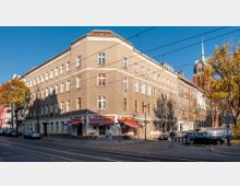 Eckgebäude in einem Wohnviertel, mit einer Bäckerei im Erdgeschoss und mehreren Fenstern an den oberen Stockwerken. Im Hintergrund ist ein Kirchturm mit grünem Dach zu sehen, umgeben von herbstlichen Bäumen und parkenden Autos.