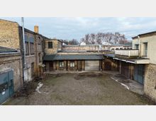 Innenhof eines alten Industrie- oder Lagerkomplexes mit Backsteinfassaden, niedrigen Anbauten und mehreren Metalltoren. Der unbefestigte Hof ist überwiegend kiesbedeckt, umgeben von Flachdächern, Rohren und einer kleinen Balkonterrasse; im Hintergrund stehen kahle Bäume.