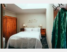 Kleines Schlafzimmer mit einem Doppelbett, weißen Kissen und einer weißen, gemusterten Bettdecke. Die Wände sind beige mit Streifenmuster, darüber eine Wanddekoration mit Schriftzug, und an den Seiten des Bettes befinden sich zwei Nachttische mit Wandlampen.