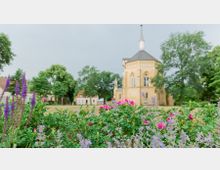 Eine historische Kirche mit hellen Außenwänden und einem kleinen Turm steht inmitten eines grünen Parks. Im Vordergrund blühen bunte Blumen, darunter rosa Rosen und violette Blüten, eingerahmt von Bäumen im Hintergrund.